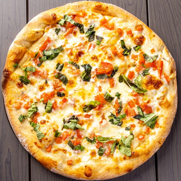 A whole round pizza with melted cheese, diced tomatoes, and fresh basil on a dark wooden table.
