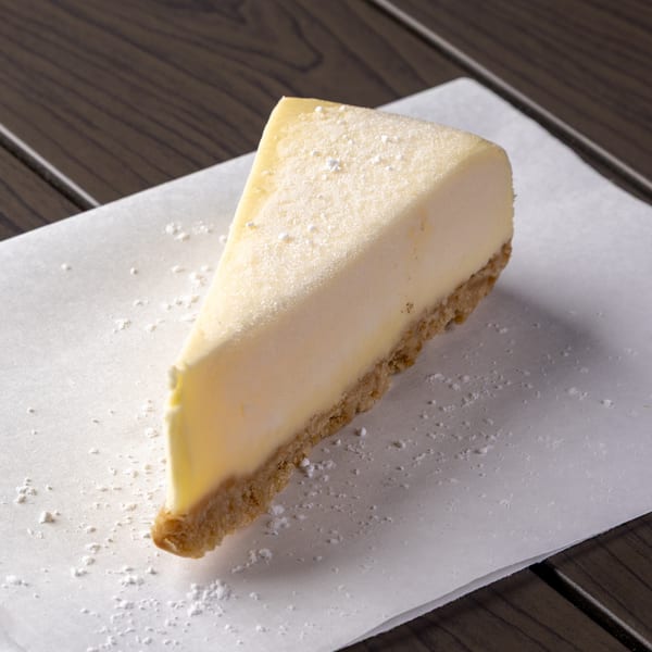 A slice of cheesecake with a graham cracker crust, dusted with powdered sugar on white parchment paper.