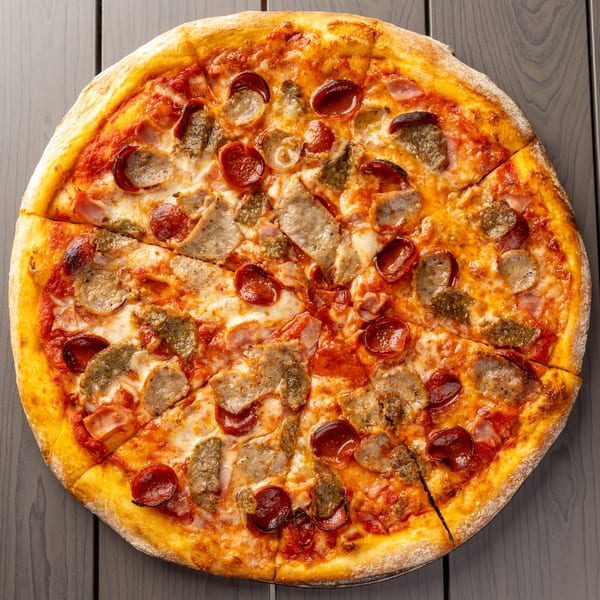 A whole meat lover's pizza, topped with pepperoni, sausage, and ham, sliced on a dark wooden table.