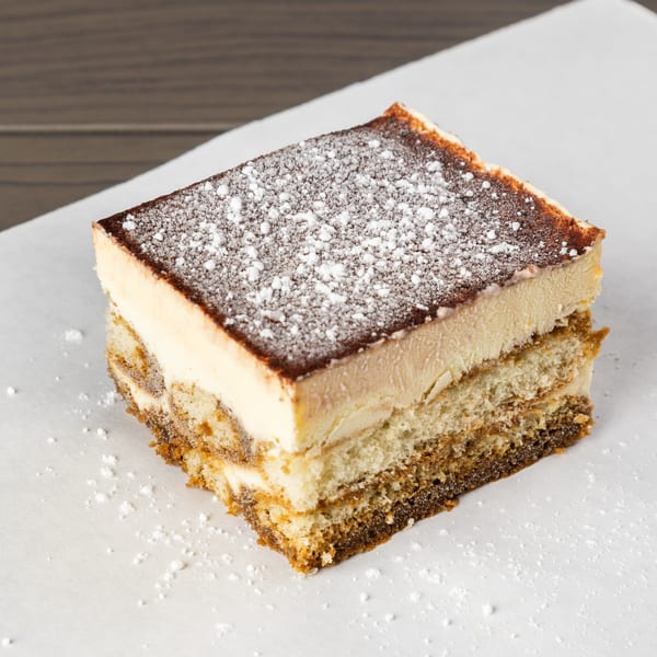 A square slice of tiramisu with layers of cake and cream, topped with cocoa and powdered sugar.