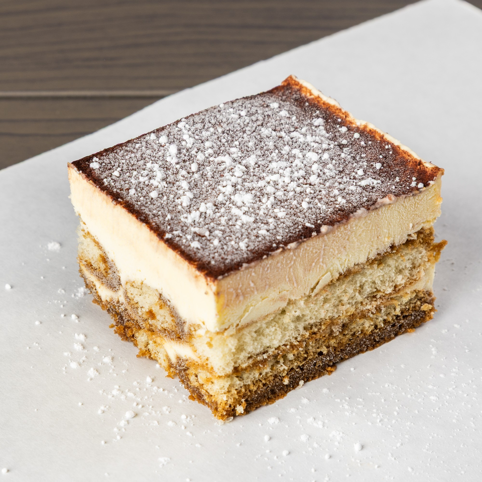 A square slice of tiramisu with layers of cake and cream, topped with cocoa and powdered sugar.