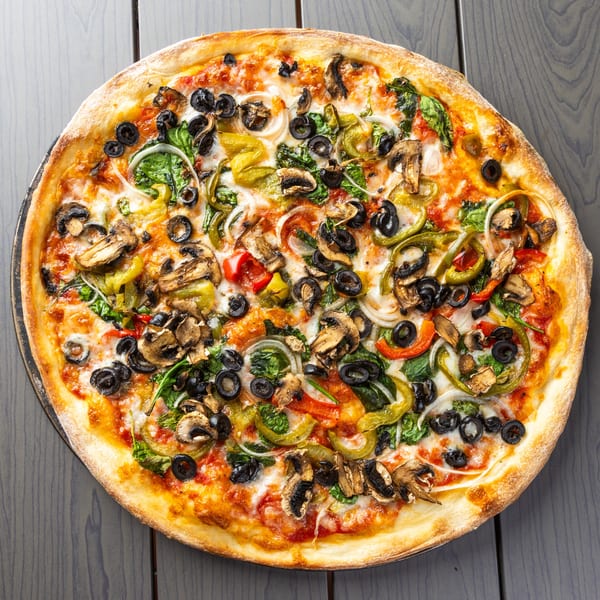 A whole vegetarian pizza with mushrooms, olives, peppers, onions, and spinach on a dark wooden table.