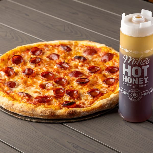 A pepperoni pizza next to a bottle of Mike's Hot Honey on a dark wooden table.