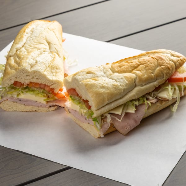 Two Annie's Pizza Subs, cut in half, filled with ham, cheese, lettuce, and tomato on a wooden table.