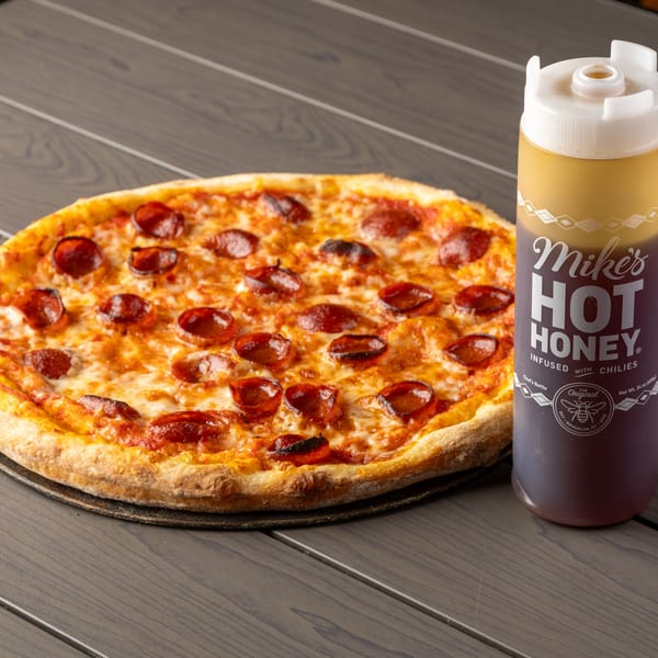 Pepperoni pizza with melted cheese next to a bottle of Mike's Hot Honey on a wooden table.
